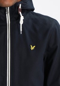 Námořnická modrá bunda s kapucí vyrobená z odolné látky, s bílým zipem a malým žlutým výšivkovým logem na levé hrudi.