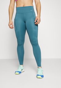 Leggings teal a vita alta con due tasche laterali, texture liscia e logo Nike. Abbinati a scarpe sportive azzurre chiare con dettagli gialli.