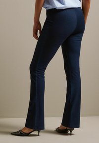 Donkerdenim flared broek met een gladde textuur, met een middelhoge taille en contrasterende stiksels. Gekozen in combinatie met zwarte puntige hakken.