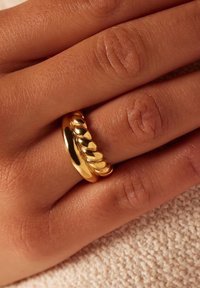 Gouden ring met een gladde band en een gestructureerd, golvend ontwerp aan één kant, rustend op een hand met subtiele huidtextuur.