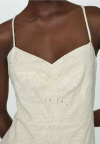 Robe crème sans manches avec broderie florale, décolleté en V et détails découpés subtils au niveau du corsage et de la taille.