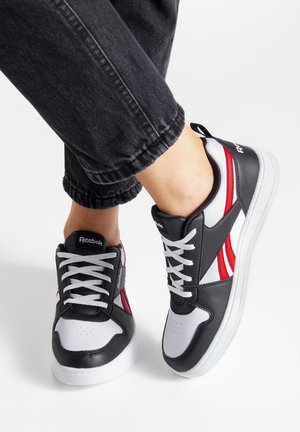 Reebok tenisky s čiernym a bielym koženým vrchom a červenými akcentmi, okrúhlou špičkou a bielymi šnúrkami, položené na bielej gumenej podrážke.