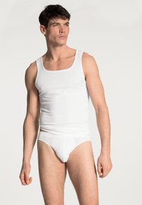 Top e slip in cotone bianco indossati da un modello maschile. Il top ha un collo a quadrato, design attillato e texture liscia. Minimalista, di colore uniforme.