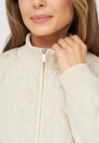 Light beige quilted jacket with a high collar and ribbed cuffs. Features a silver zipper and diamond pattern detailing.
