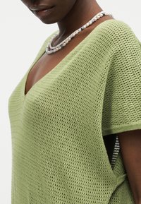 Γυναίκα που φοράει ανοιχτό πράσινο πλεκτό τοπ με V-λαιμόκοψη και περιδέραιο με πέρλες και μπλε χάντρες, εμφανιζόμενη από τους ώμους μέχρι το μέσο του σώματος.