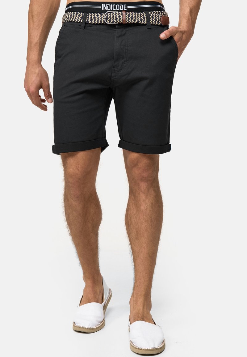 Homme portant un short noir à ourlet roulé, ceinture tissée, chaussures blanches sans lacets et taille INDICODE visible, une main dans la poche.