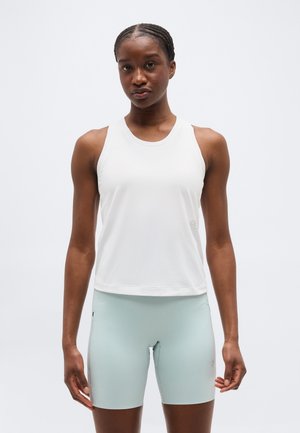 Femeie atletă purtând un top alb fără mâneci și pantaloni scurți de bicicletă cu talie înaltă, de culoare albastru deschis, stând în picioare pe un fundal gri simplu.