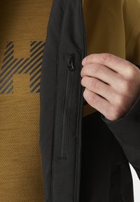 Schwarze Jacke mit einer Reißverschlusstasche und einer kontrastierenden braunen Innenlage mit strukturiertem Muster. Hand, die den Reißverschluss zieht.