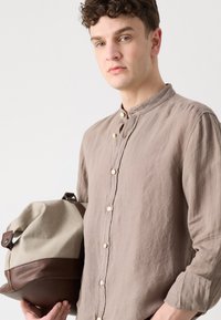 Linnen overhemd in lichtbruin met een bandkraag en houten knopen. Houdt een canvas tas vast met een brown leren basis en accenten.