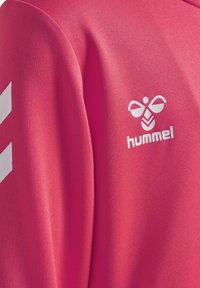 Różowa koszulka sportowa Hummel z teksturowanym materiałem. Posiada biały logo i akcenty w kształcie chevronu na lewym ramieniu.