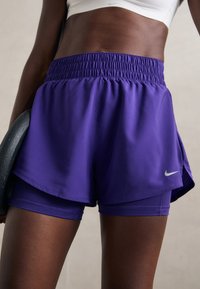 Idrettslig person iført lila lagdelte shorts og hvit crop-top, som holder en svart vektplate ved siden av seg.