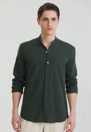 Ung mand iført mørkegrøn skjorte med mandarin-krave med ærmerne rullet op og lyse bukser, står mod en ensfarvet baggrund.