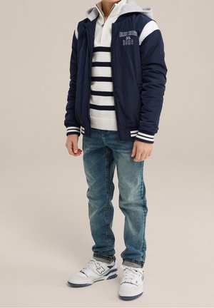 Chaqueta de piloto de la marina con acentos blancos, sudadera gris claro debajo, suéter a rayas, vaqueros azules y zapatillas blancas con detalles azules.