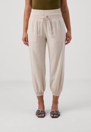 Personne portant un pantalon beige à taille et poignets élastiques avec poches avant et des sandales noires à bout ouvert sur un fond clair uni.