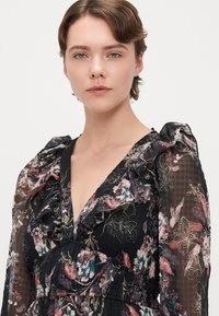 Čierne kvetinové šaty s volánovými, priehľadnými rukávmi a hlbokým výstrihom do V. Majú farebné botanické vzory a štruktúrovanú látku.