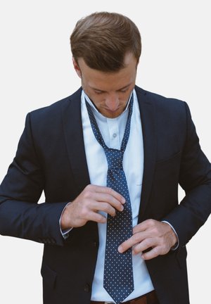 Une cravate à pois navy ajustée par une personne en costume noir, portant une chemise blanche et une ceinture brune visible. Texture lisse, forme élancée.