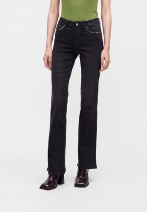 Frau trägt schwarze Straight-Leg-Jeans mit Nieten an den Taschen, kombiniert mit dunklen Stiefeln mit Absatz und einem grünen ärmellosen Oberteil.