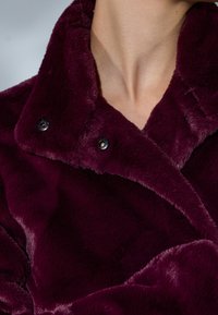 Burgundy keinoturkki pehmeällä pinnalla, suuressa kauluksessa ja kahdessa mustassa nappikiinnityksessä. Ominaisuutta on tyylikäs ja valahtava muotoilu.