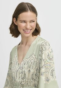 Rochie verde deschis cu decolteu adânc în V, având un design cu model în nuanțe de bej, negru și culori pastelate. Material moale și fluid, cu mâneci căzute.