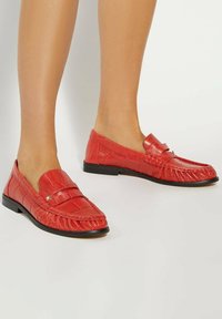 Red faux leather loafers with crocodile texture, slip-on design, and black rubber soles. Features decorative stitching and a front strap.