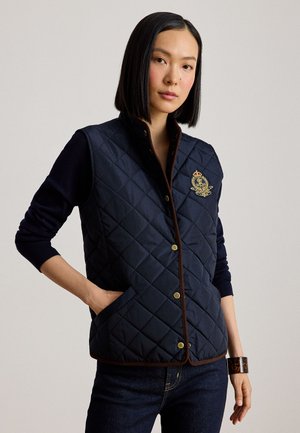 Donna con capelli neri lisci che indossa un gilet trapuntato blu navy con stemma ricamato, una maglia scura a maniche lunghe, jeans e un bracciale a polsino in tartaruga.