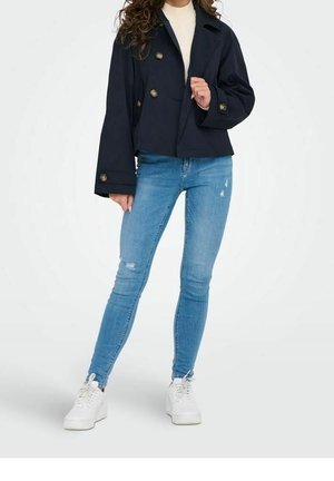 Vrouw die een marineblauw dubbelgeknoopt jasje draagt, een witte top, lichtblauwe gescheurde skinny jeans en witte sneakers, staand tegen een effen achtergrond.
