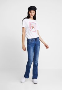 Kvinne iført en svart caps, hvit t-skjorte med grafikk, blå jeans og hvite joggesko som står mot en ensfarget hvit bakgrunn.