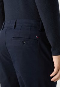 Tamno plave hlače s gumbom na stražnjem džepu, ravnim prednjim dizajnom i teksturiranim materijalom. Imaju mali detalj logotipa u crvenoj, bijeloj i plavoj boji.