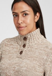 Beige strikkegenser med høy, ribbestrikket krage, som har tre brune knapper på stykke. Teksturen er tykk og myk.