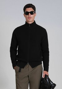 Pull en maille noire zippé avec col montant, présentant un ourlet fendu et un tissu légèrement texturé. Porté avec des lunettes de soleil sombres et un pantalon beige.
