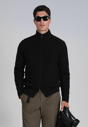 Black ribbed zip-up sweater with high collar, featuring a split hem and slightly textured fabric. Worn with dark sunglasses and beige pants.