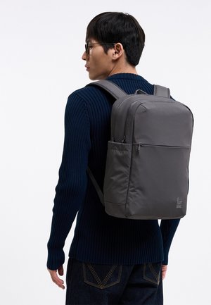 Hombre con gafas que lleva un suéter azul marino y vaqueros oscuros, que lleva una mochila grande gris con bolsillos delanteros y laterales con cremallera.