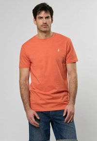 Tričko s krátkým rukávem a kulatým výstřihem v korálové barvě z bavlny. Na hrudi je malé bílé logo. V kombinaci s modrými džínovými šortkami.