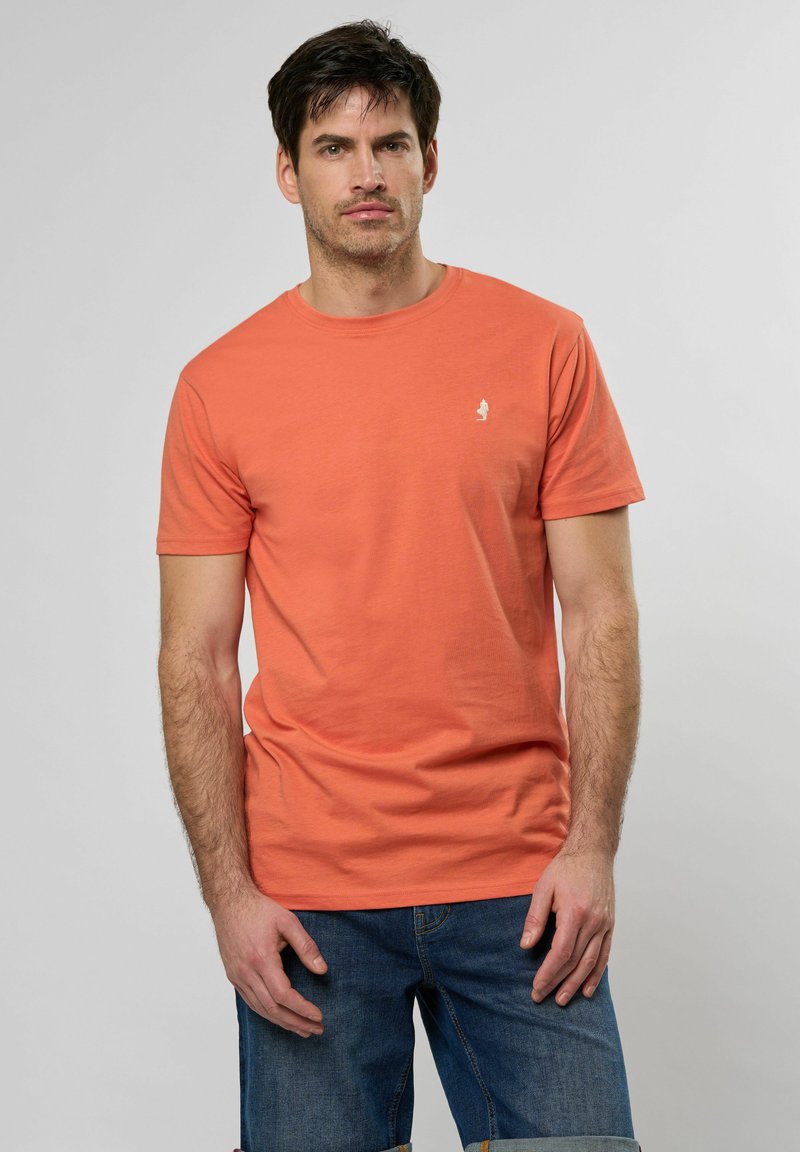 Tričko s krátkým rukávem a kulatým výstřihem v korálové barvě z bavlny. Na hrudi je malé bílé logo. V kombinaci s modrými džínovými šortkami.