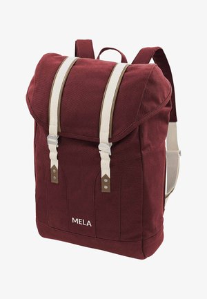 Baardige man in een wit t-shirt, blauwe spijkerbroek en zwarte sneakers draagt een grote rode rugzak met het etiket "MELA" over één schouder.