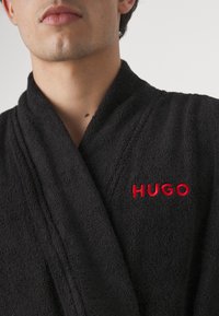 Peignoir de bain en terry noir avec un col châle et un logo "HUGO" brodé en rouge sur la poitrine. Texture douce avec un design classique.