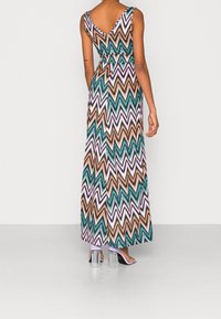 Robe maxi avec un motif en chevrons en teal, orange et rose. Présente un dos en V et une taille cintrée, fabriquée en tissu léger. Talons transparents visibles.