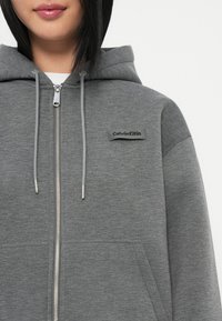 Pilkas užtrauktukas su gobtuvu, sidabrinis užtrauktukas ir juodas Calvin Klein logotipo ženklas ant kairės krūtinės. Minkšta audinio tekstūra.