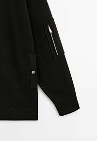 Vêtement noir à manches longues avec une poche zippée sur le haut du bras et une poche à bouton-pression près de la taille sur un fond uni.
