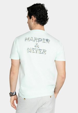 Mann mit lockigem dunklem Haar, der ein mintgrünes T-Shirt mit dem Schriftzug „HARDER & NIXER“ in dekorativen Buchstaben auf dem Rücken trägt, kombiniert mit hell gestreiften Hosen.