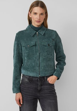 Veste en velours côtelé bleu canard avec un col, fermeture zip et deux poches poitrine. Surface texturée et coupe courte. Associée à un jean foncé.