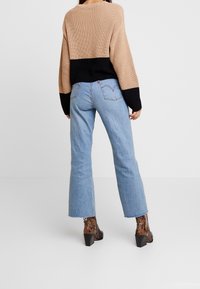 Femme portant un pull beige et noir, un jean droit bleu clair et des bottes à talons imprimé serpent marron, debout sur un fond blanc.