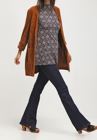 Hnědý semišový dlouhý kardigan přes vzorované tričko, kombinovaný s tmavými rozšířenými džíny a černými dřevěnými dřeváky s podpatkem. Minimální doplňky.