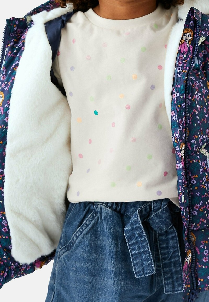 Enfant portant une veste à motif floral doublée en polaire blanche, un pull crème à pois colorés, et un pantalon en denim bleu avec une ceinture nouée.