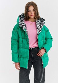 Žena oblečená v zelenej zateplenej bunde so zebra vzorom vo vnútri, svetloružovom svetri, čiernych džínsoch a zlatom náhrdelníku, stojí proti bielej pozadí.