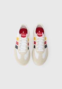 Hvide og beige sneakers lavet af ruskind og mesh-materialer. Har røde, gule og sorte striber, hvide snørebånd og rød inderside.