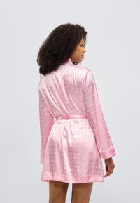 Ροζ σατέν ρόμπα με μακριά μανίκια, γιακά και ζώνη δετή στη μέση. Διαθέτει διακριτικό μοτίβο σε όλη την επιφάνεια, απαλό ύφασμα και μήκος μέχρι τα μέσα μηρού.