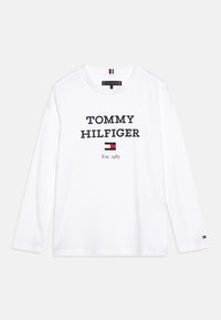 Bílá košile s dlouhým rukávem, na které je tučně napsáno "TOMMY HILFIGER" v černém písmu. Logo obsahuje malý detail červeno-bílé-modré vlajky na rukávu.