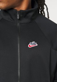 Close-up van een persoon in een zwart Nike zip-up jack met een rood en wit logo op de linkerborst.