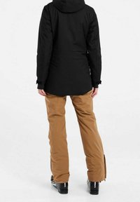 Personne portant une veste noire, un pantalon de neige beige et des chaussures de ski noires, debout de dos sur un fond blanc.